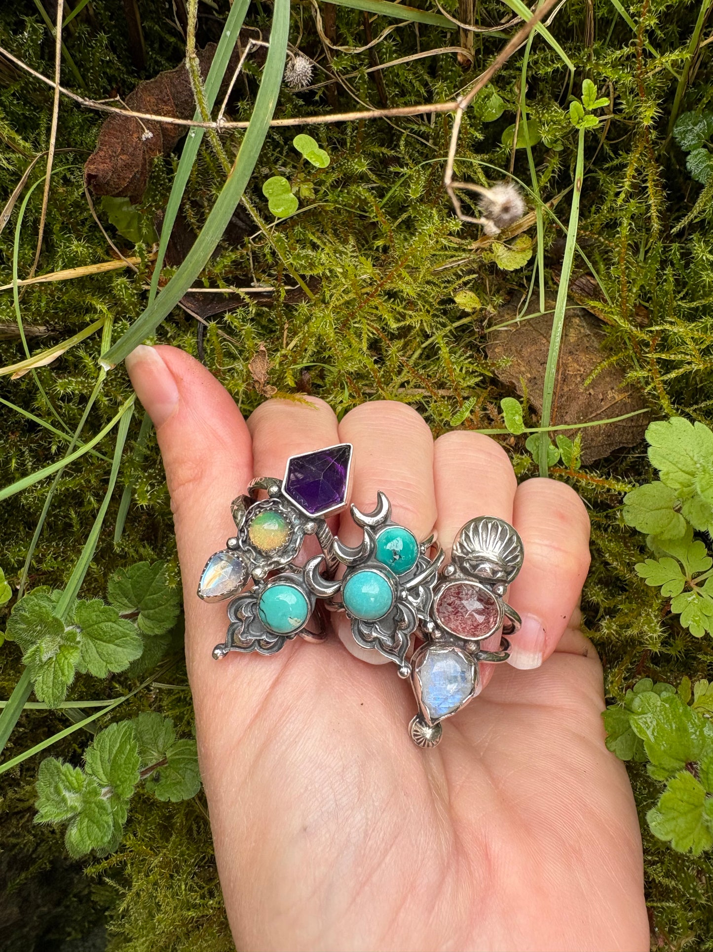 Hand holding multiple handmade decorative ring with gemstones against a natural mossy background