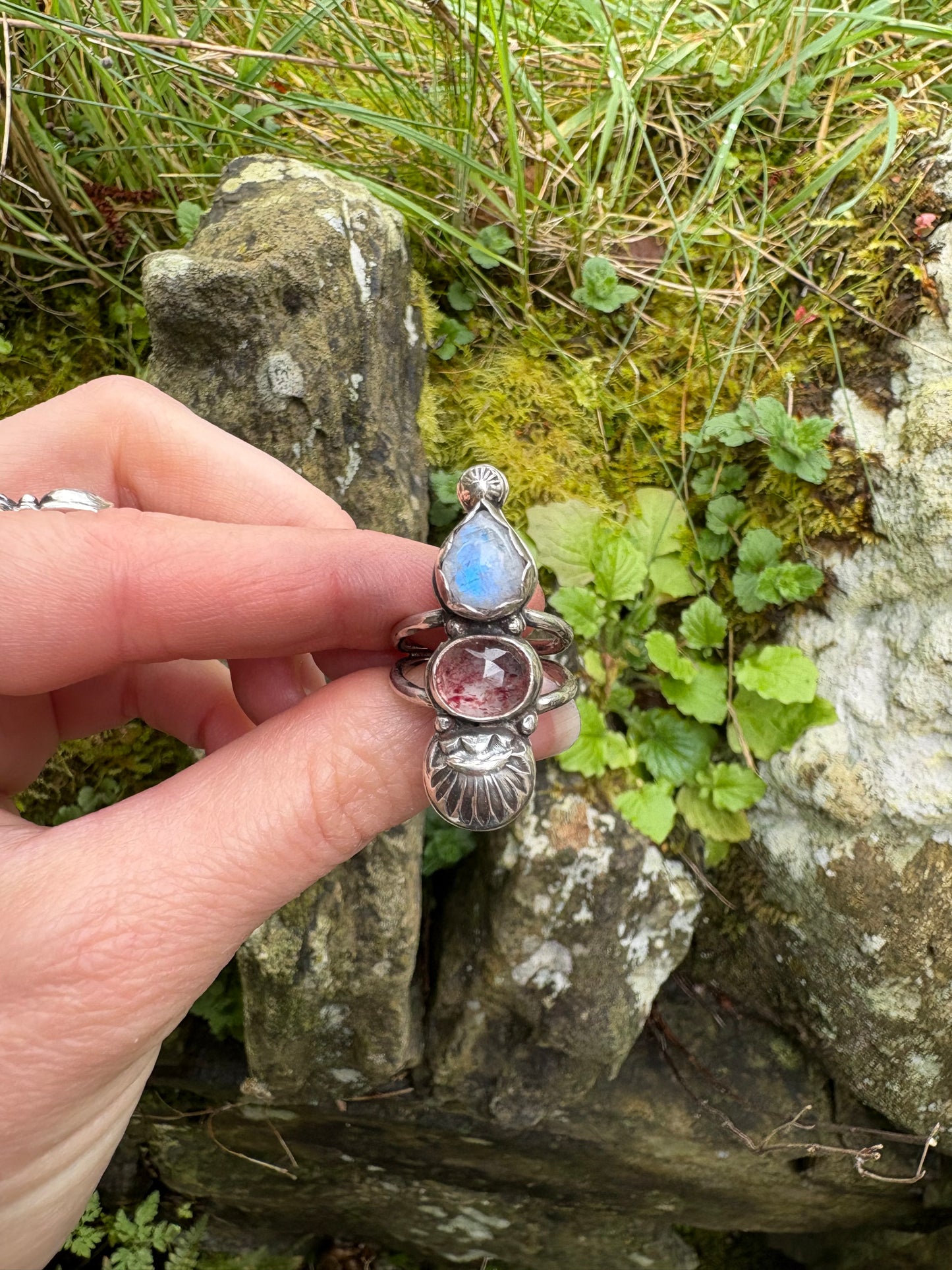 Hand holding a sterling silver handmade ring with gemstones , moonstone, strawberry quartz, against a natural background of rocks and grass.
