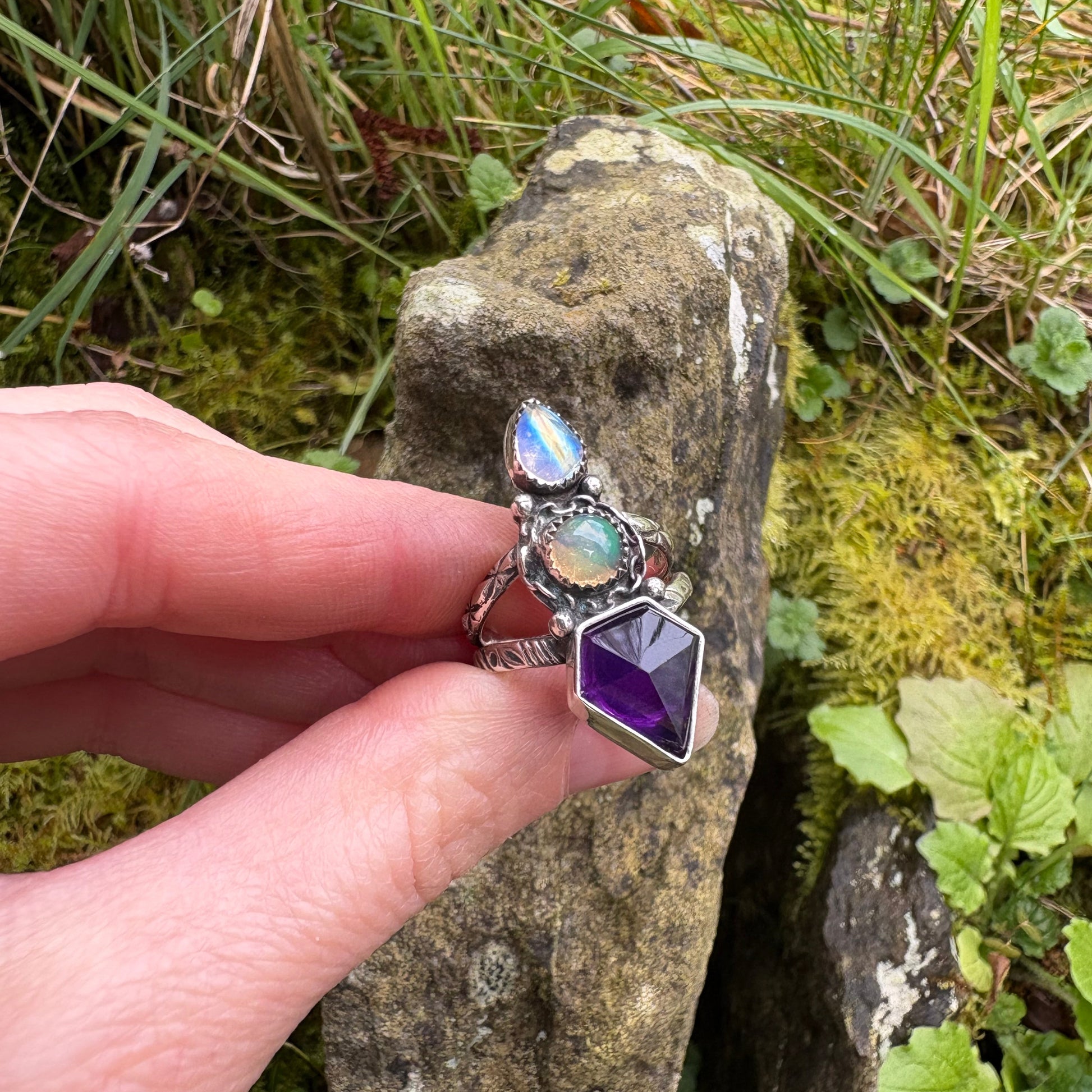 hand holding handmade ring of amethyst opal and moonstone against mossy background of rocks and leaves 