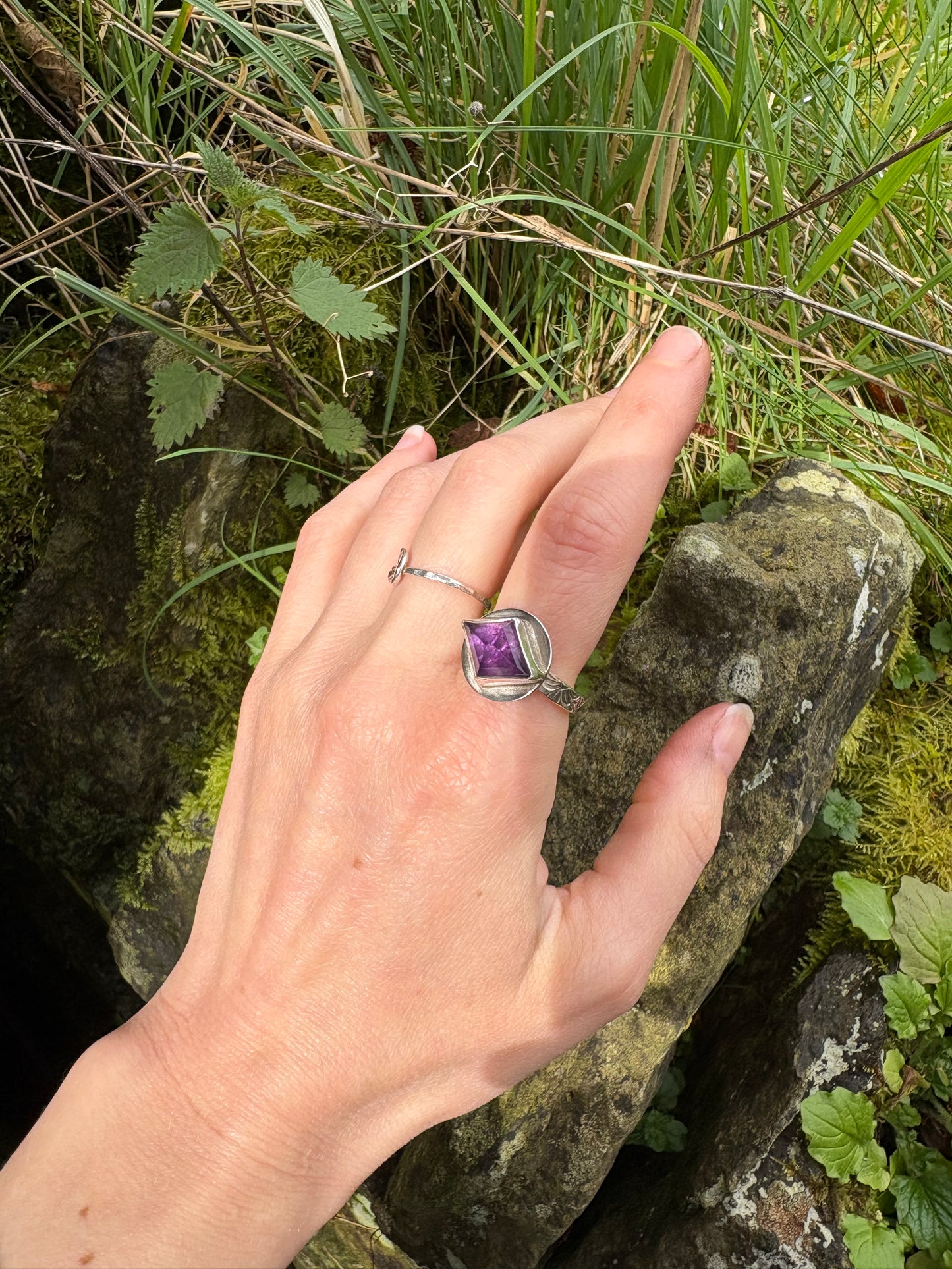 Hand wearing a silver ring with a purple gemstone against a natural background