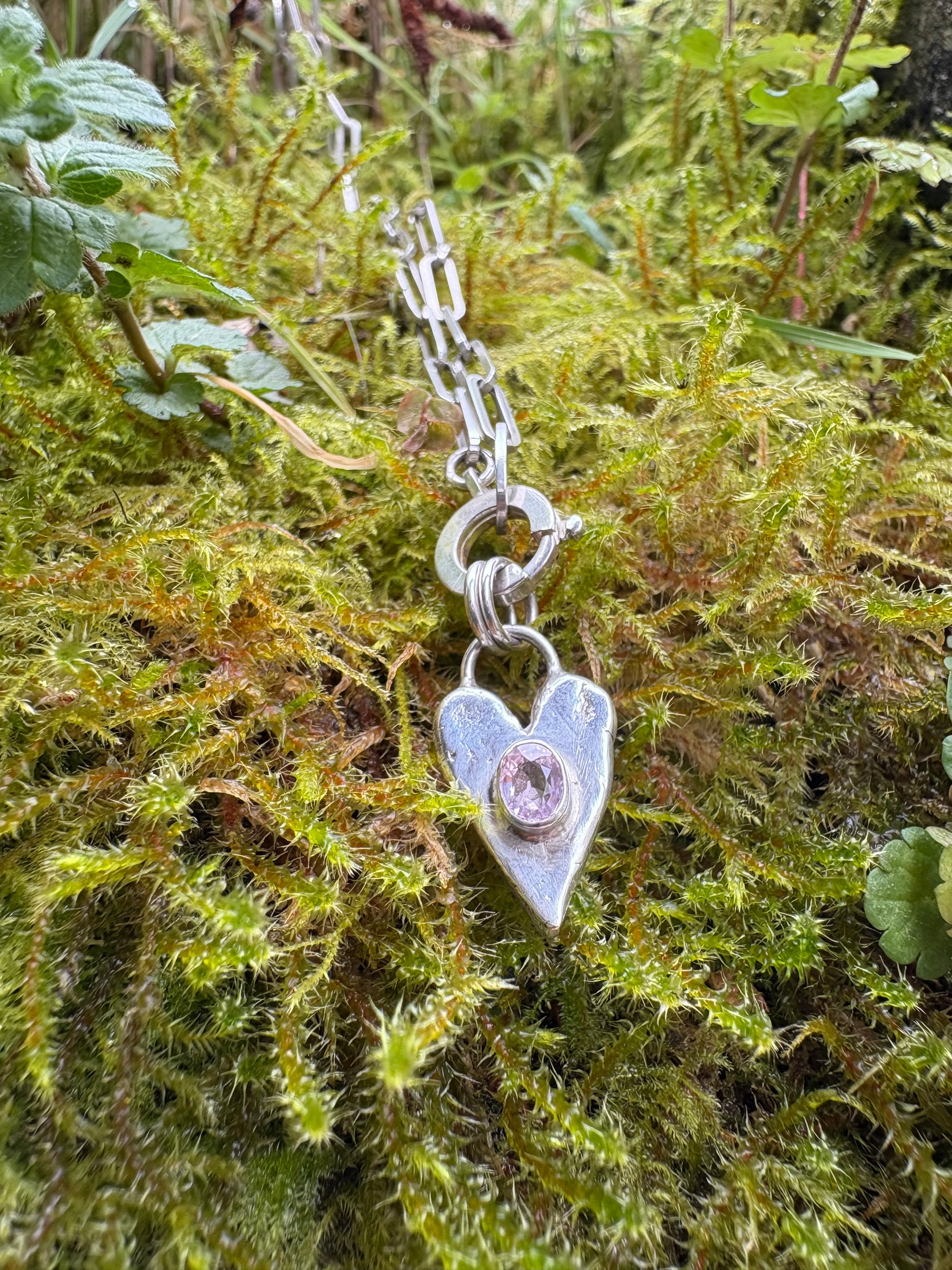 Silver heart-shaped charm with pink tourmaline set in the middle, on an 18" chain lying on green moss